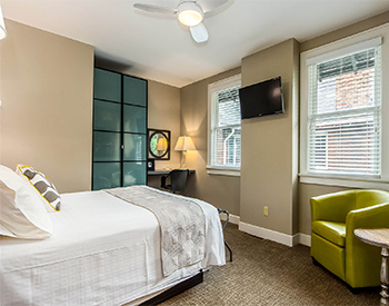 Bed with TV mounted on the wall and green chair under a widow.