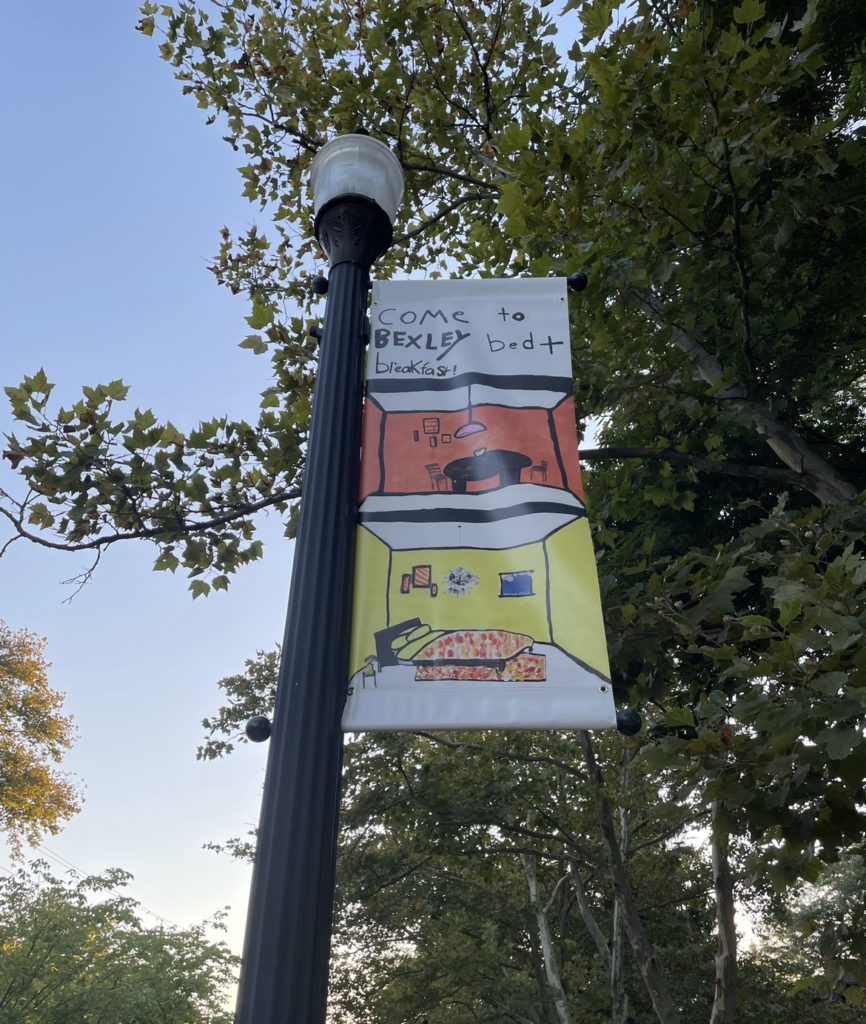 Looking up at a street lamp with a sign made by a child that says "Come to Bexley Bed + Breakfast." The sign shows a guest room and the dining room. A tree dominates most of the foreground while a blue evening sky filters through the leaves.