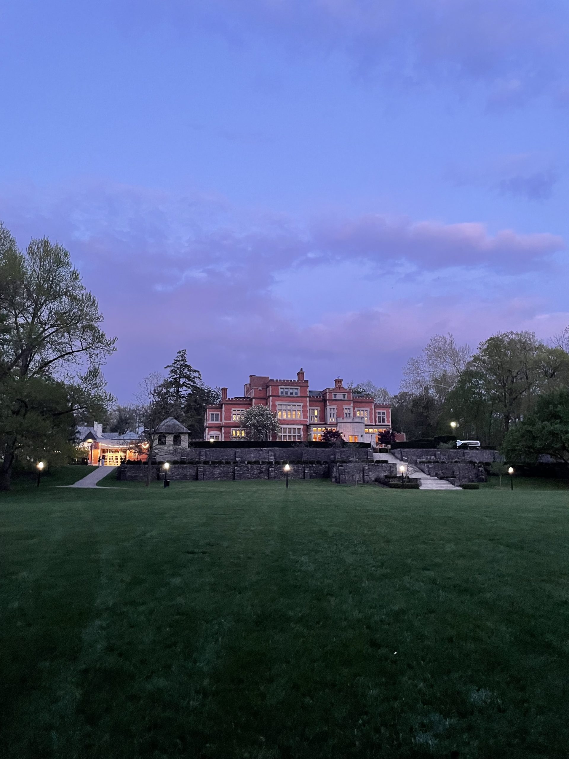 A brick mansion sits on a dark green grassy hill under a sunset sky of blue, purple, and pink clouds.