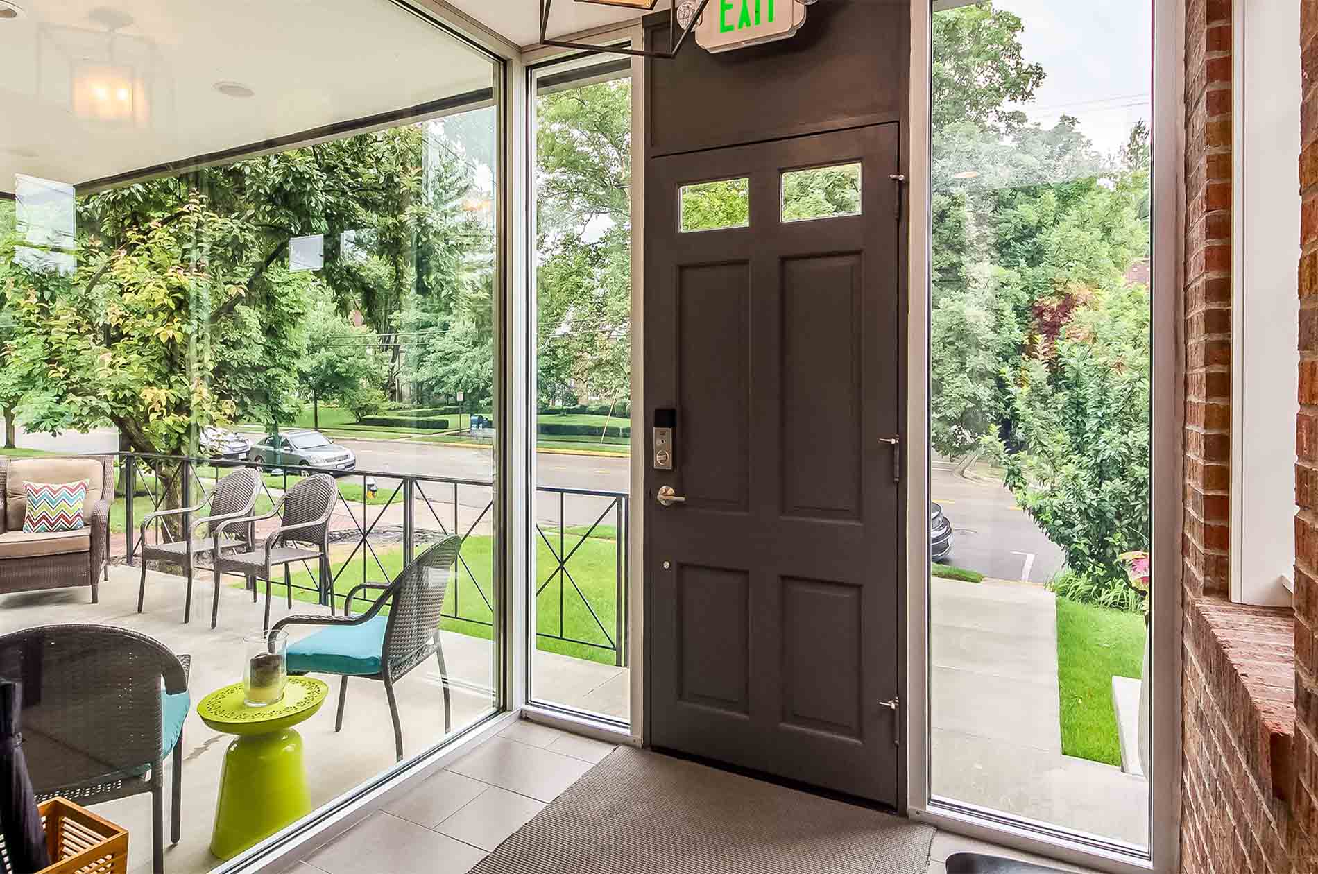 Glass entry from inside looking outside upon an exterior sitting area surrounded by green trees, and cars parked on the street.