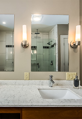 Bathroom counter with multiple mirrors and sink. In mirror, glass shower with multiple showerheads visible.