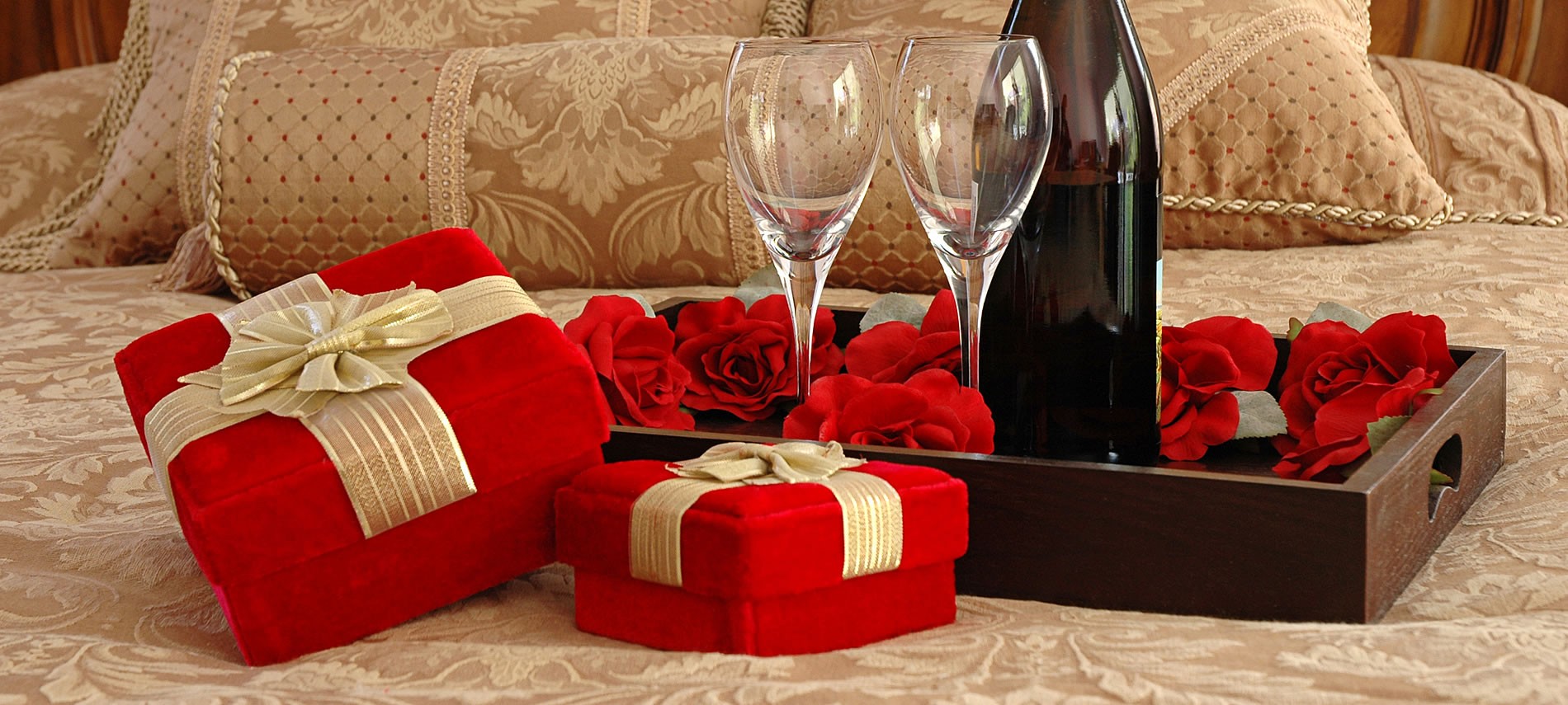 Red velvet boxes with decorative bows next to a dark wooded bed tray with wine, wine glasses and red roses, all set upon a tan bed.