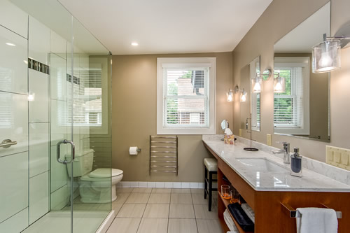 Long view into bathroom showcasing modern appointments. Lamps upon the wall above sink, and glass shower opposite.