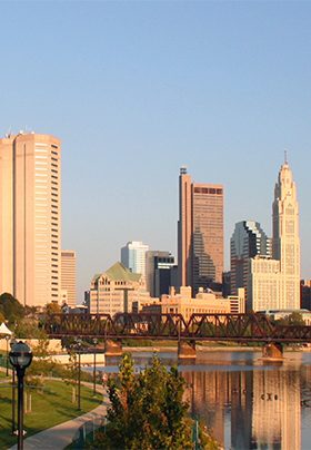 Skyline View of city in background. Foreground is green grass, with ducks on water with a girder bridge crossing the waterway.